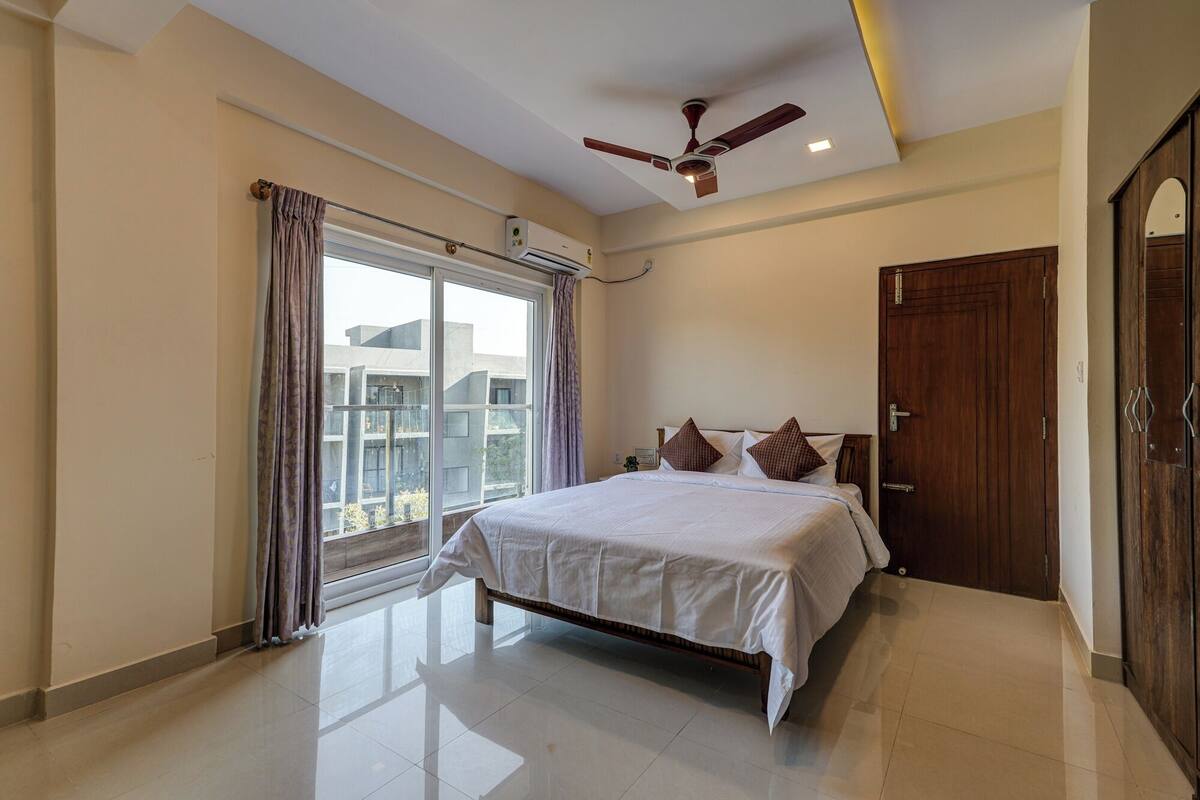 Quarto com uma cama, um ventilador de teto, janela com cortinas e uma porta.
