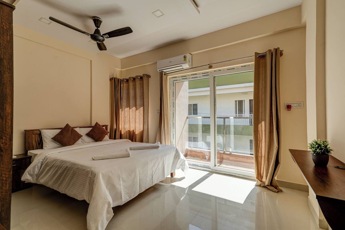 Quarto com cama, cortinas, ventilador de teto, aparelho de ar condicionado e vista por uma porta de vidro.