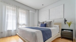 Een moderne slaapkamer met een groot bed, een nachtkastje met een lamp en een raam met transparante gordijnen.
