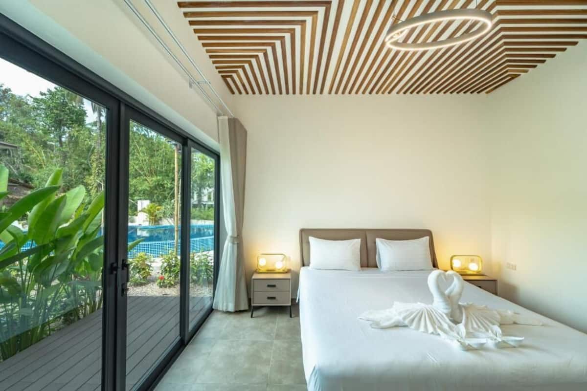 A modern hotel room with a large bed, two bedside tables with lamps, a ceiling with a geometric pattern, and a sliding glass door leading to a pool area.