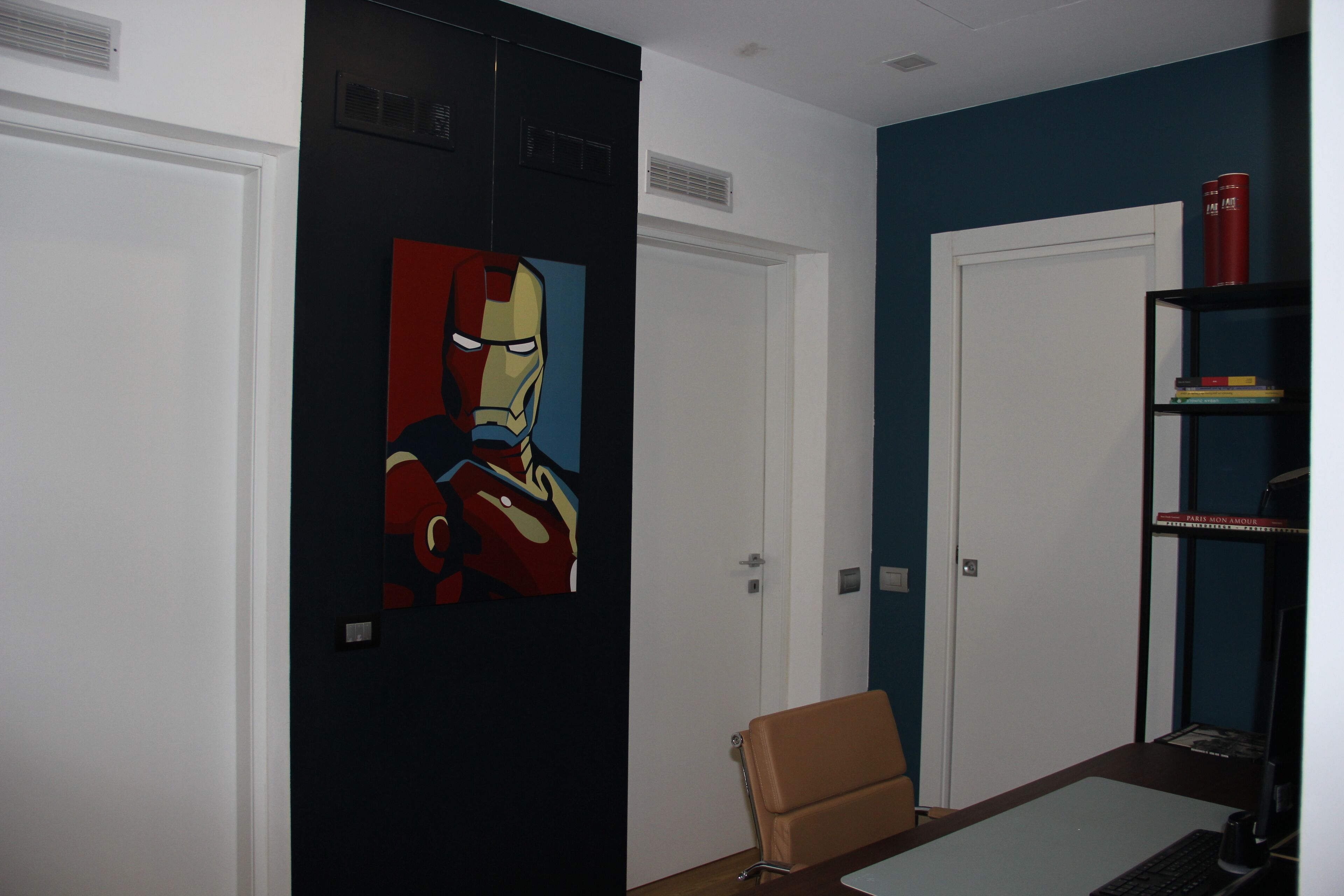 Una habitación con una puerta, una silla marrón, un escritorio con una computadora y una estantería con libros y una lata roja.