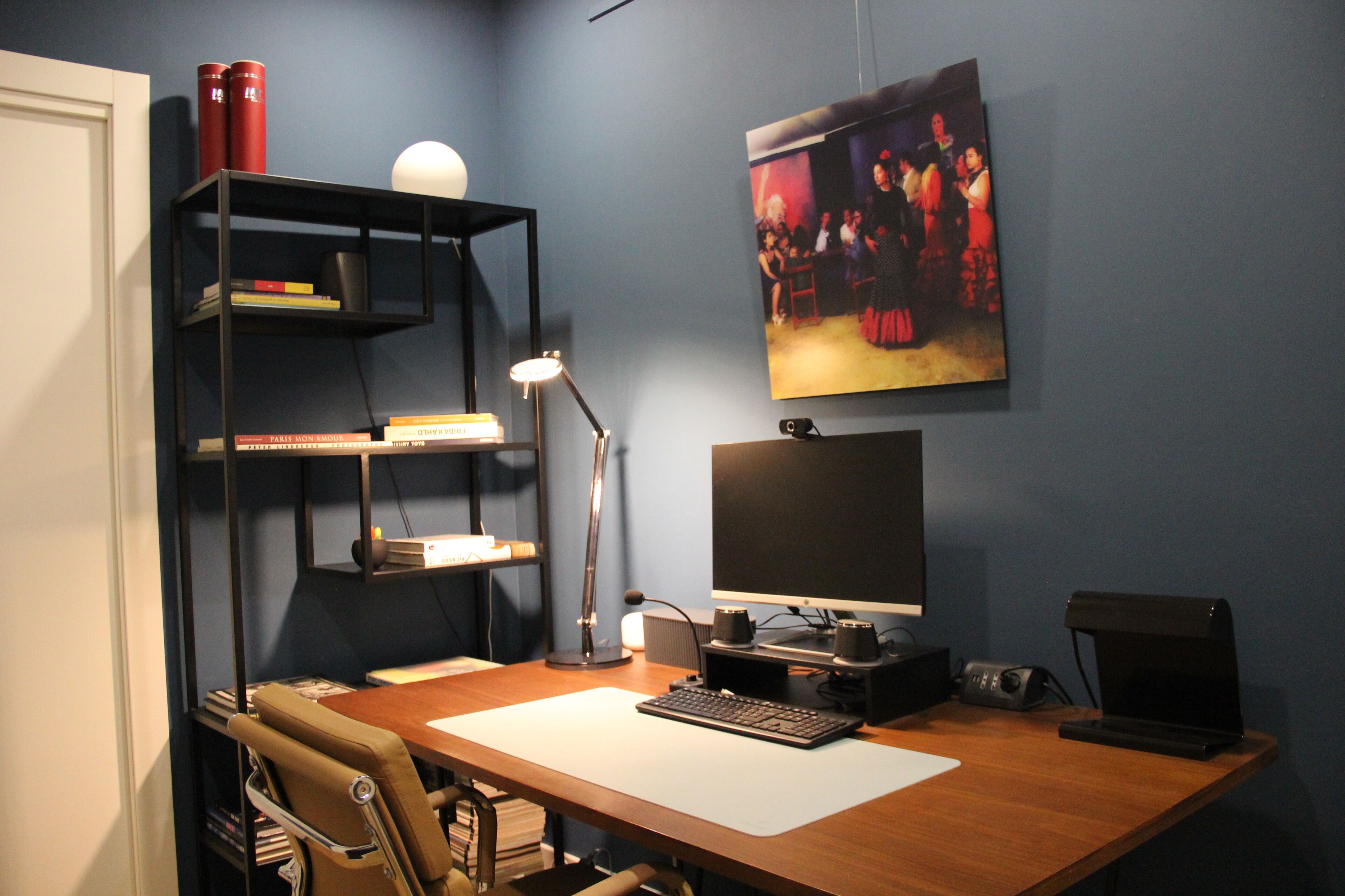 A home office setup with a desk, computer, shelves, and a painting.