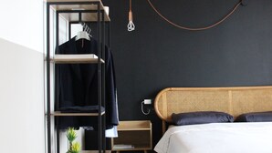 Una camera da letto con soffitto in legno, un letto con biancheria bianca, uno sgabello, una mensola con vestiti e una pianta in vaso.