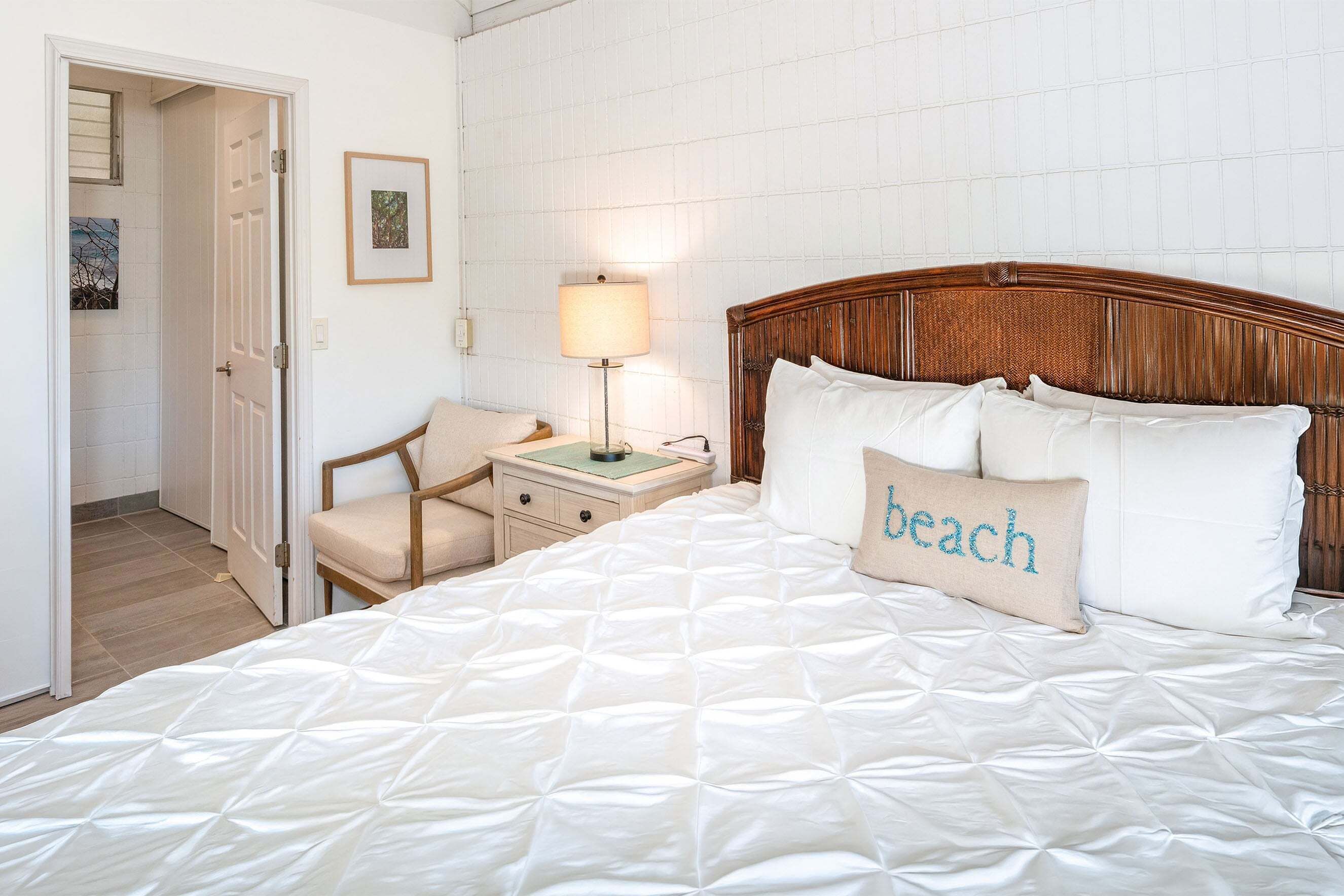 Una cama bien hecha con cubrecama acolchado, una lámpara de noche, una silla y una puerta que da a otra habitación.