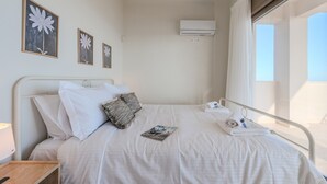 Un llit ben fet amb llençols blancs, una almoçada, una revista i una tovallola. Hi ha quadres emmarcats a la paret, un aparell d'aire condicionat i una finestra amb cortines.
