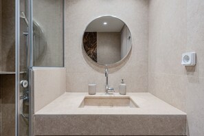 Un baño moderno con cabina de ducha de vidrio, lavamanos con resguardo de mármol y espejo redondo.