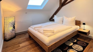A bedroom with a skylight, wooden beams, a bed with white linens, a bedside table with a lamp, and a decorative floor rug. - Rebenblick Ferienwohnung (Landau in der Pfalz)