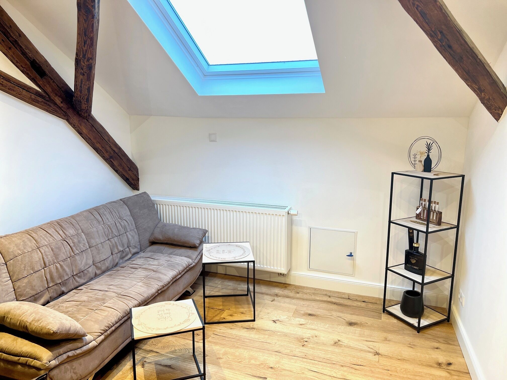 Ein Loft-Zimmer mit schrägverdachter Decke, Holzbalken, einem weißen Heizkörper, einem schwarzen Metallregal und einem beigefarbenen Sofa.