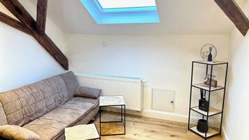 Ein Loft-Zimmer mit schrägverdachter Decke, Holzbalken, einem weißen Heizkörper, einem schwarzen Metallregal und einem beigefarbenen Sofa.