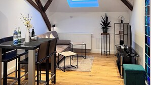 A loft-style room with a sloped ceiling, exposed wooden beams, a skylight, a dining area with a table and chairs, a sofa, a small side table, a radiator, a potted plant, a black shelving unit, and a green ottoman. - Rebenblick Ferienwohnung (Landau in der Pfalz)