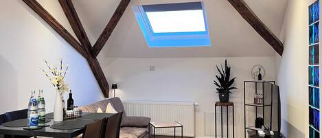 Une pièce de style « loft », avec un plafond en pente, des poutres apparentes, un puits de lumière, un coin repas avec une table et des chaises, un canapé, une petite table d’appoint, un radiateur, une plante en pot, une étagère noire et un pouf vert.