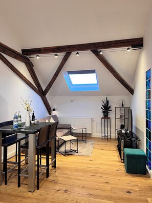 A loft-style room with a sloped ceiling, exposed wooden beams, a skylight, a dining area with a table and chairs, a sofa, a small side table, a radiator, a potted plant, a black shelving unit, and a green ottoman. - Rebenblick Ferienwohnung (Landau in der Pfalz)