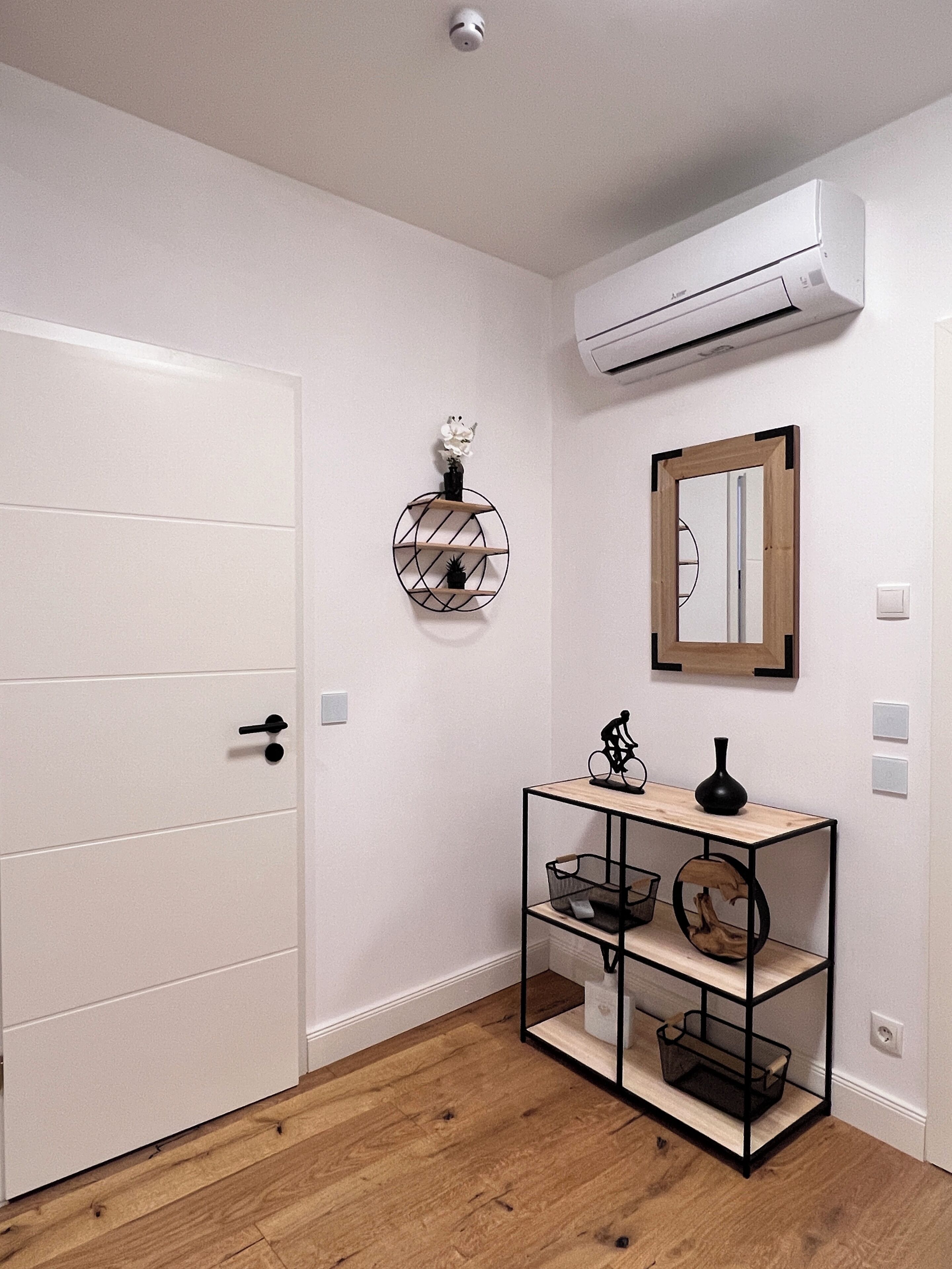 A hallway with a white door, a wall-mounted air conditioner, a mirror, and a shelf with decorative items.