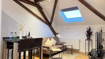 A loft-style room with exposed wooden beams, a skylight, a sofa, a dining table with chairs, and a small side table with a plant.