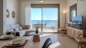 Una sala moderna con sofá, mesa de comedor y sillas, un balcón con vista al mar y un televisor sobre un mueble.