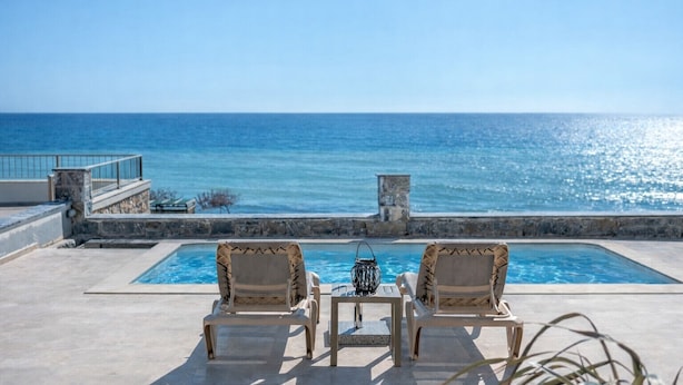 Zona junto a la piscina con dos sillones de descanso y una mesa, con vista al mar azul y cristalino.