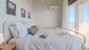 Una cama bien hecha con sábanas blancas, una almohada, una revista y una toalla. En la pared hay cuadros enmarcados, un equipo de aire acondicionado y una ventana con cortinas.