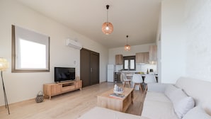 A modern living room with a sofa, coffee table, TV, and dining area with chairs and a kitchen in the background. - Kedros Villa Tessera (Viannos)