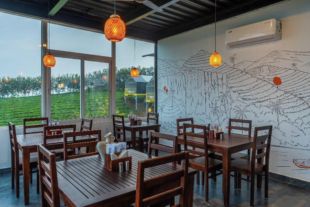 A restaurant with wooden tables and chairs, hanging pendant lights, and a mural of a landscape on the wall.