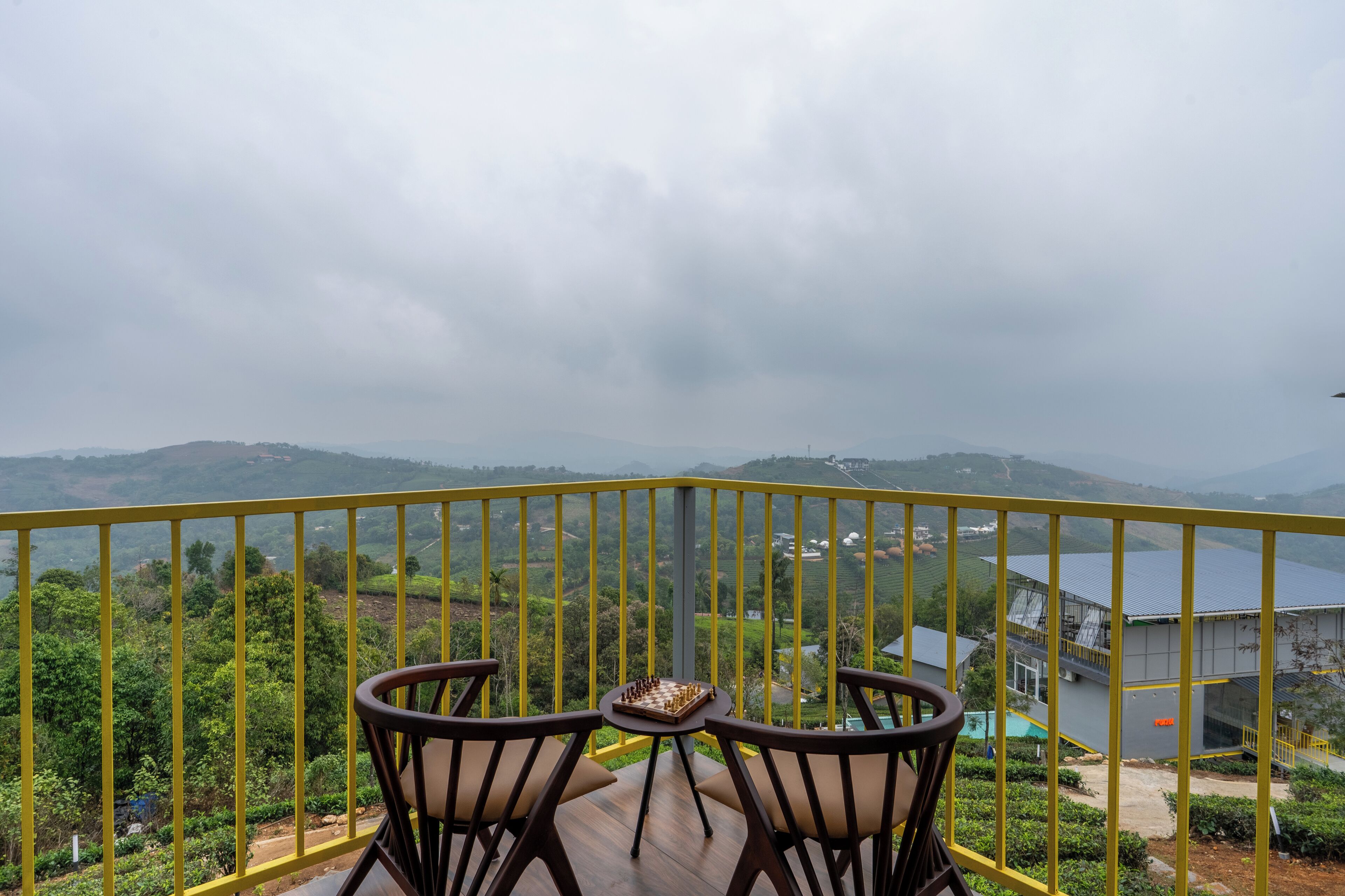Een balkon met een schaakbord, twee stoelen en uitzicht op glooiende groene heuvels en gebouwen.
