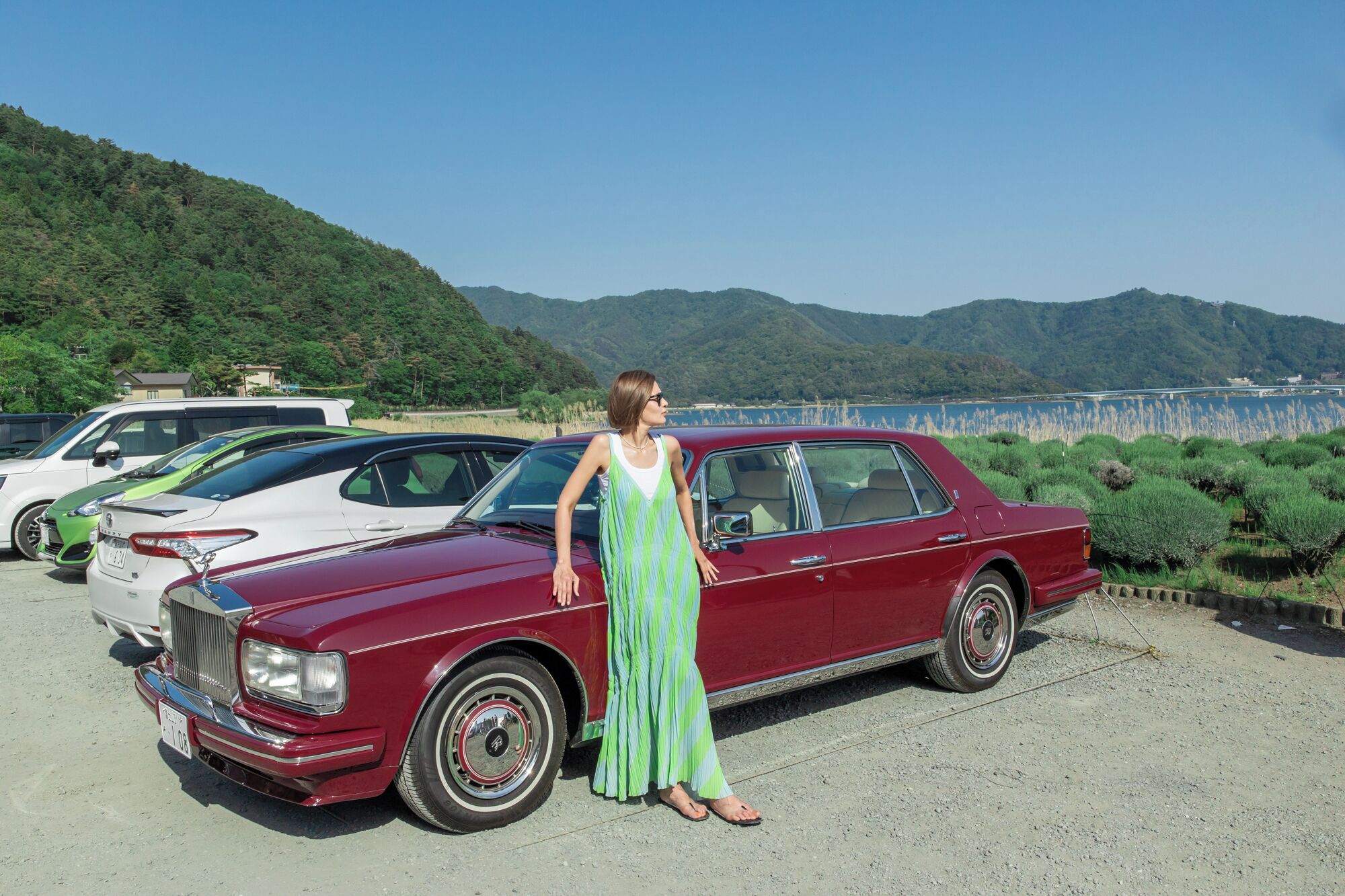 A vintage red car parked in a lot with a woman standing beside it, surrounded by greenery and hills in the background.