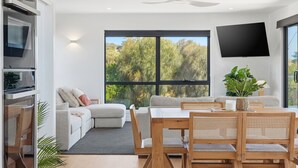 Una sala de estar moderna con sofá, una mesa de comedor y sillas. Se aprecia un ventanal con vistas a árboles.