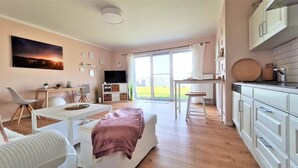 Ein modernes Wohnzimmer mit einer Couch, einem Esstisch, einem Fernseher und einer Küchenzeile mit Schränken und einem Fenster.