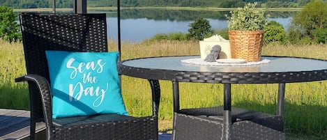 A wicker chair with a pillow, a small round table with a plant, and a view of a lake and forest.