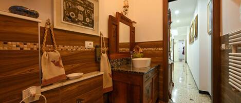 Un baño con tocador de madera, lavamanos y espejo. Hay un perchero con toallas y un cuenco decorativo en la encimera.