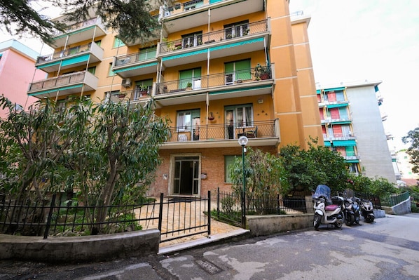 Um edifício residencial de vários andares com varandas, portão fechado e scooters estacionadas.