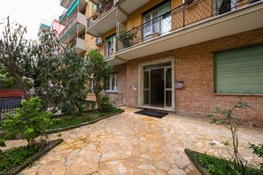 Um edifício residencial de vários andares com um pátio pavimentado em pedra, varandas com grades e uma janela com persianas verdes.