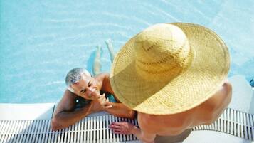 Eine Person liegt mit einem Strohhut auf einer Liege am Pool, eine andere sitzt am Poolrand.