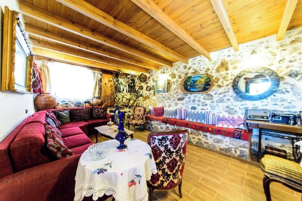 A cozy living room with a stone wall, wooden ceiling beams, a red sofa, a patterned armchair, and a small table with a vase.