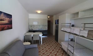 Habitación compacta con literas, una kitchenette pequeña, una mesa de comedor con sillas y un sofá.