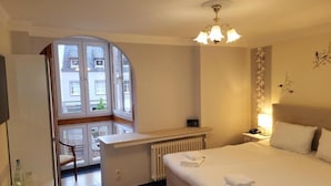 A hotel room with a bed, a desk, a chair, a radiator, a window, and a chandelier. - Hotel am Markt - Restaurant Meridiana (Saarburg)