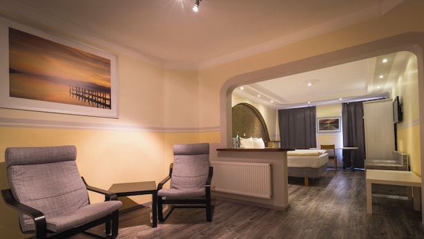 A modern hotel room with a wooden floor, two grey armchairs, a small table, a bed, a mirror, and a painting on the wall. - Hotel am Markt - Restaurant Meridiana (Saarburg)