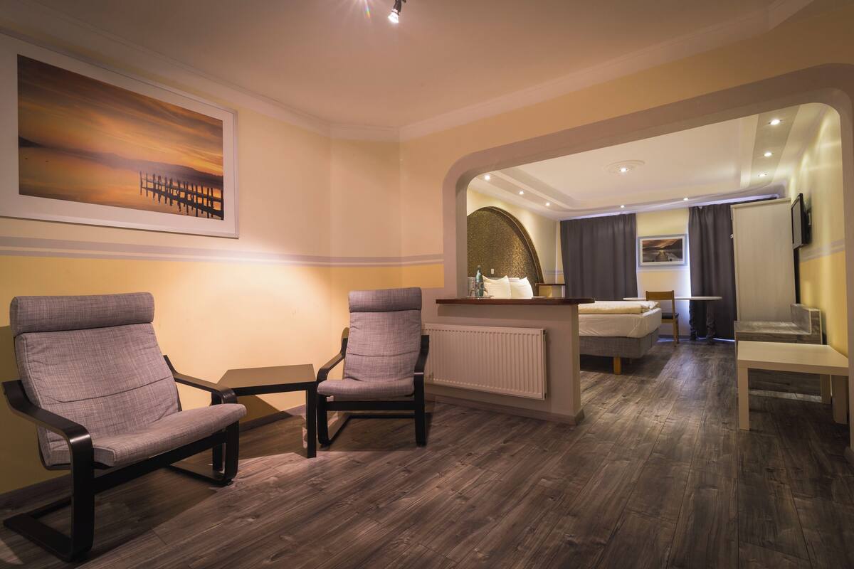A modern hotel room with a wooden floor, two grey armchairs, a small table, a bed, a mirror, and a painting on the wall.