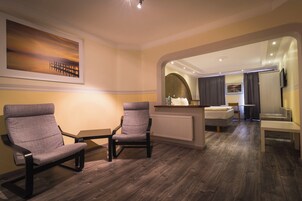 A modern hotel room with a wooden floor, two grey armchairs, a small table, a bed, a mirror, and a painting on the wall.