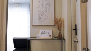 Ein Empfangsbereich mit einem gerahmten Kunstwerk über einem Willkommensschild, einem kleinen Tisch mit einer Vase getrockneter Pflanzen und einem schwarzen Stuhl.
