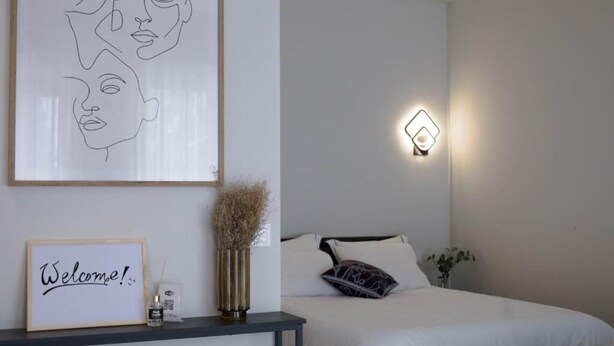 Quarto com cama, mesa de cabeceira com um letreiro de boas-vindas emoldurado, luminária na parede e uma obra de arte na parede.