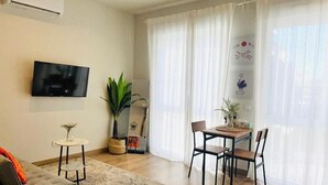 Uma sala de estar com televisão de tela plana, um sofá, uma mesa de jantar com cadeiras, uma pequena mesa lateral e uma janela grande com cortinas leves.