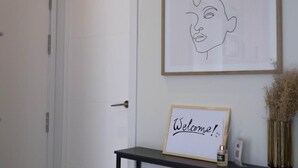 Uma porta branca com puxador prateado, um letreiro de boas-vindas emoldurado e um vaso com plantas secas sobre uma mesa preta.