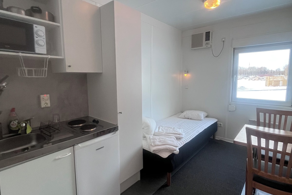 A compact kitchenette with a microwave, stove, and sink. A single bed with a mattress and pillows is visible, and a wooden chair is placed near a window.