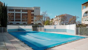 Una piscina con cubierta azul, rodeada por una cerca blanca, y contigua a un edificio con varios balcones.