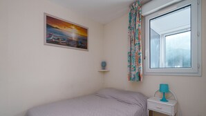 Un dormitorio con cama, ventana con cortinas, un cuadro en la pared y una mesita de noche con lámpara.