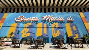 Un murale con il testo 'Santa Maria di Leuca', area salotto all'aperto con sedie e tavoli in vimini e un tendone a righe.