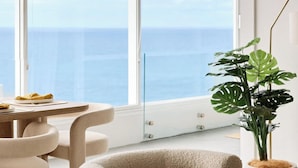 En moderne, rund, beige stol med en træsokkel, placeret på en tekstureret beige måtte i et rum med store vinduer med udsigt over vand.