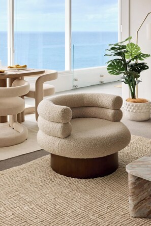 A modern, round, beige chair with a wooden base, positioned on a textured beige rug in a room with large windows overlooking a body of water. - Cliffside Escape w/ Pool & Panoramic Ocean Views (Vaucluse)