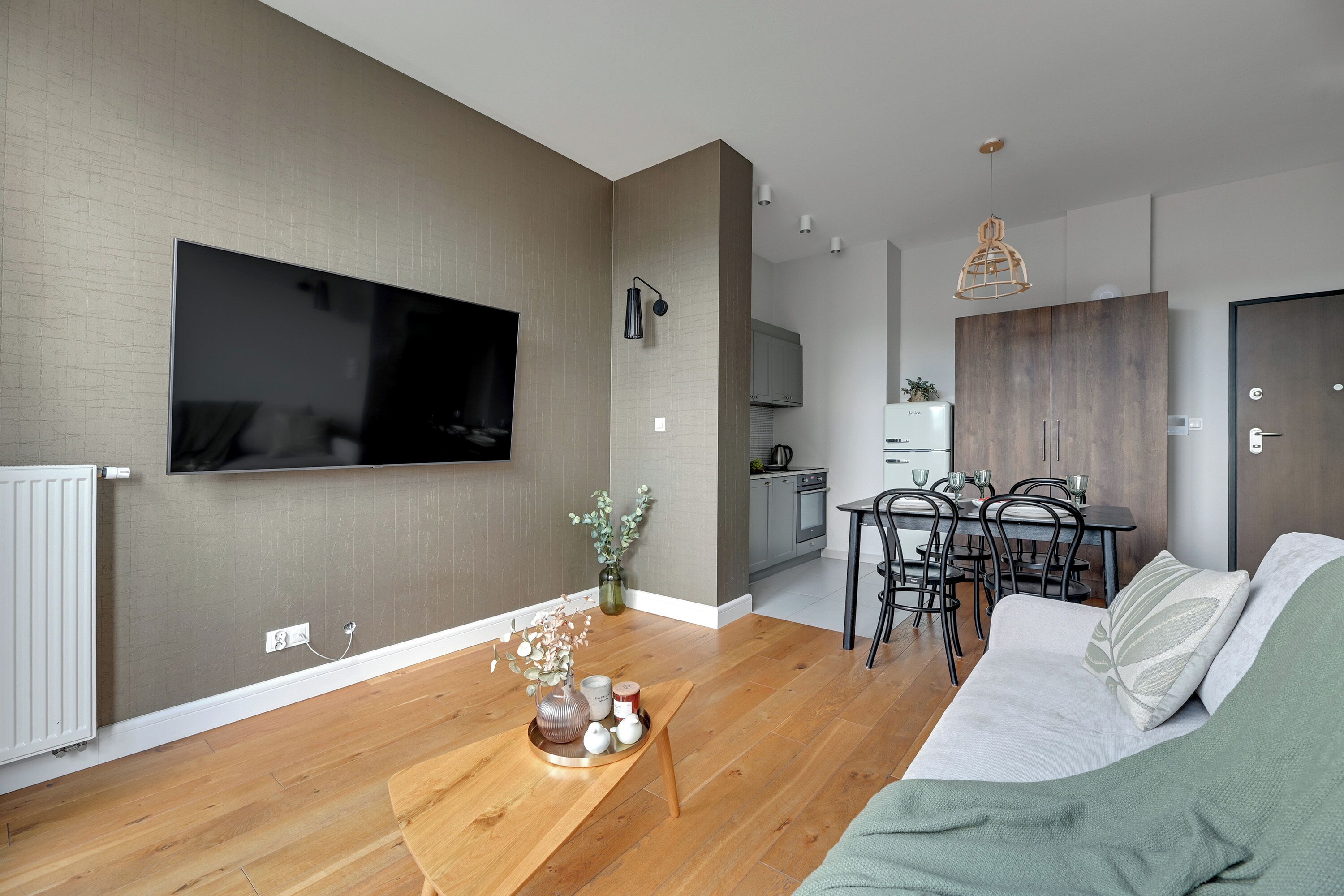 A modern living room with a flat-screen TV, wooden flooring, a dining area with a table and chairs, and a kitchen in the background.