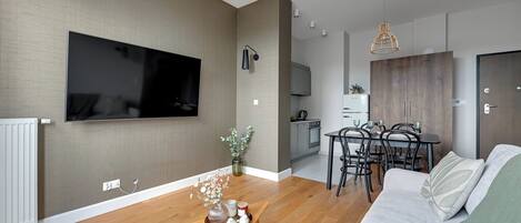 A modern living room with a flat-screen TV, wooden flooring, a dining area with a table and chairs, and a kitchen in the background.
